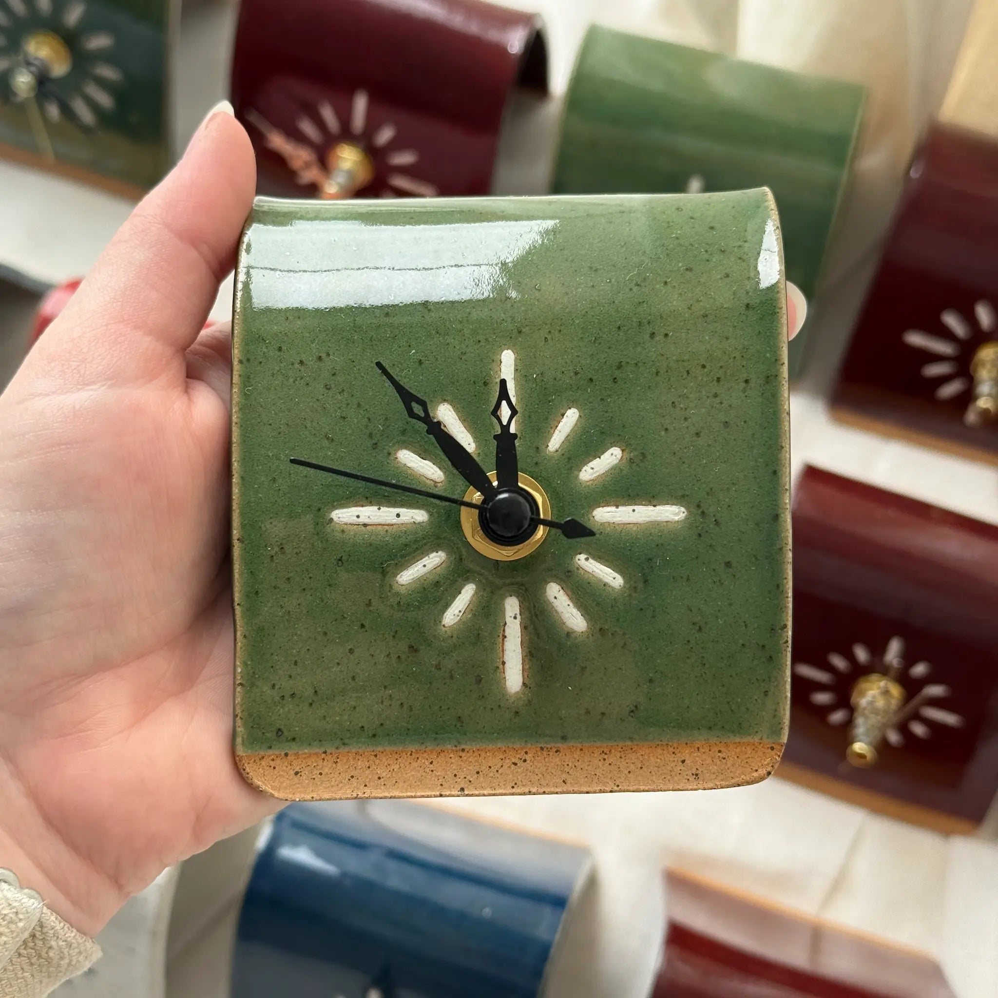 Hand holding a small green ceramic clock with black hands against a blurred background of similar items.