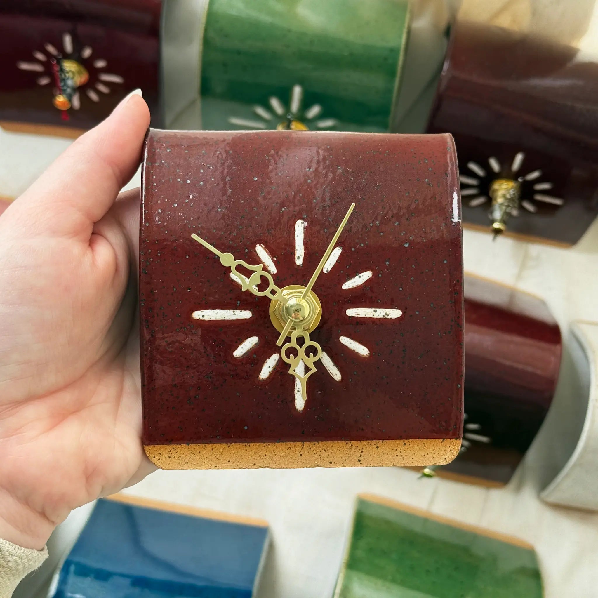 Hand holding a small square cordovan clock with gold hands against a blurred background of other clocks.