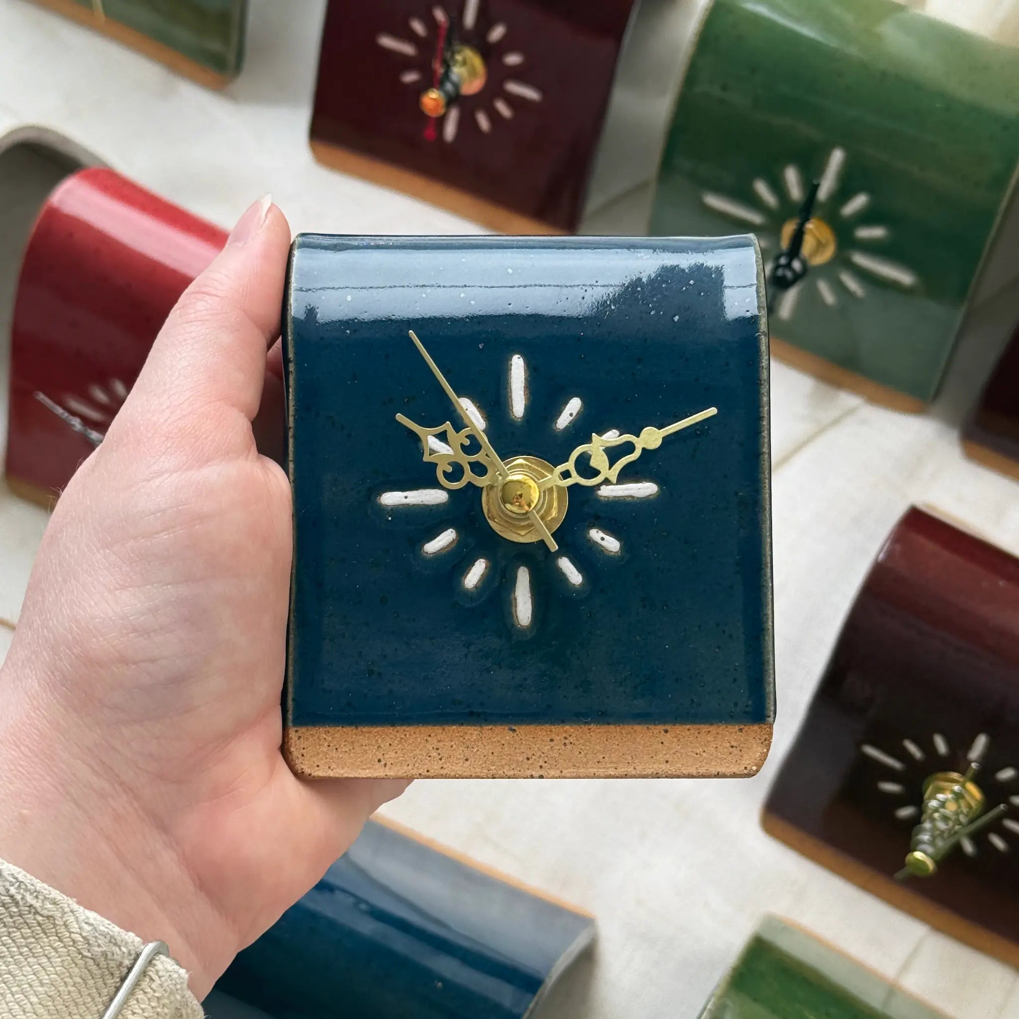 Hand holding a small dark blue square clock with gold hands, surrounded by other similar clocks in the background.