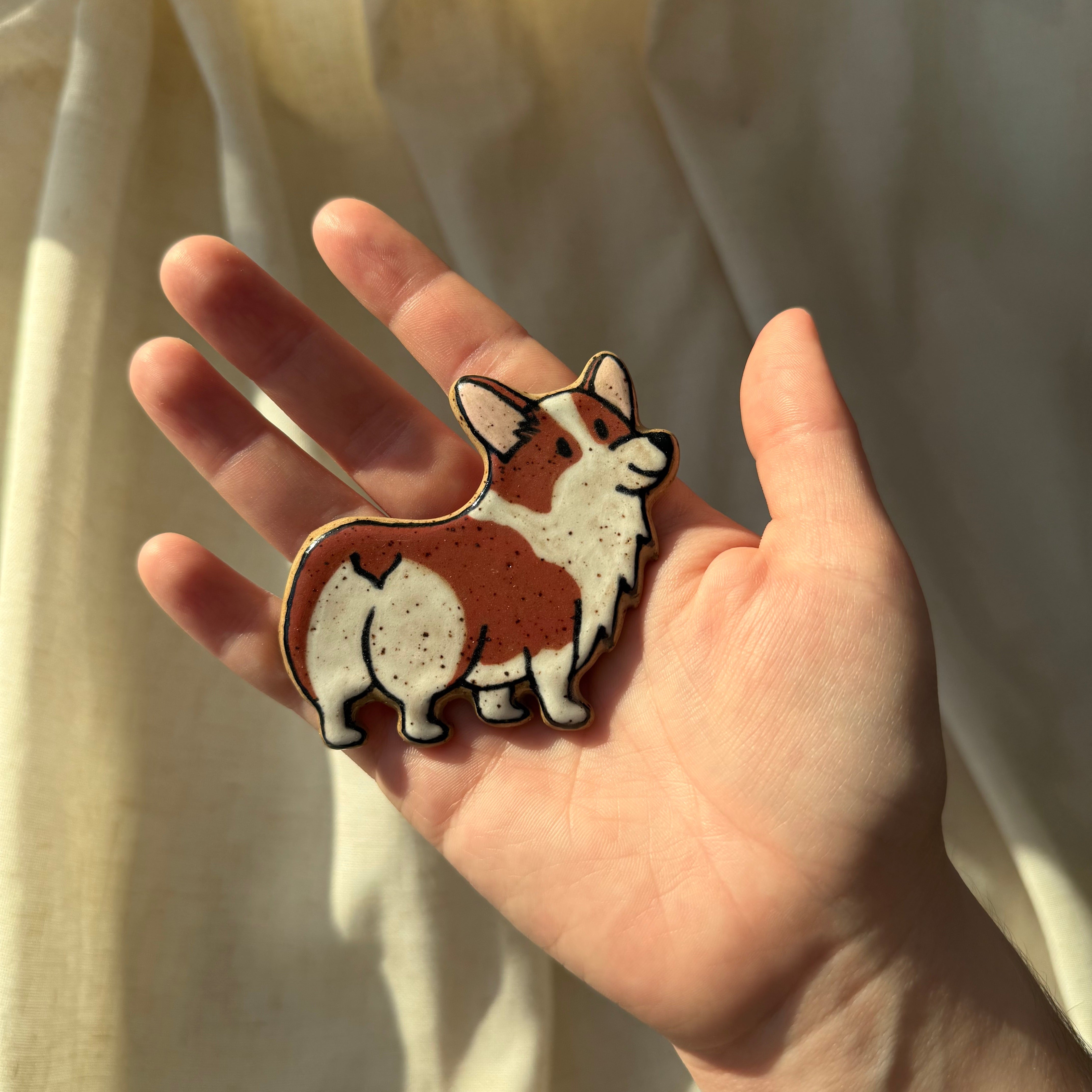 A hand holding a small ceramic magnet shaped and handpainted like a corgi dog, with a soft cream fabric backdrop in warm sunlight.