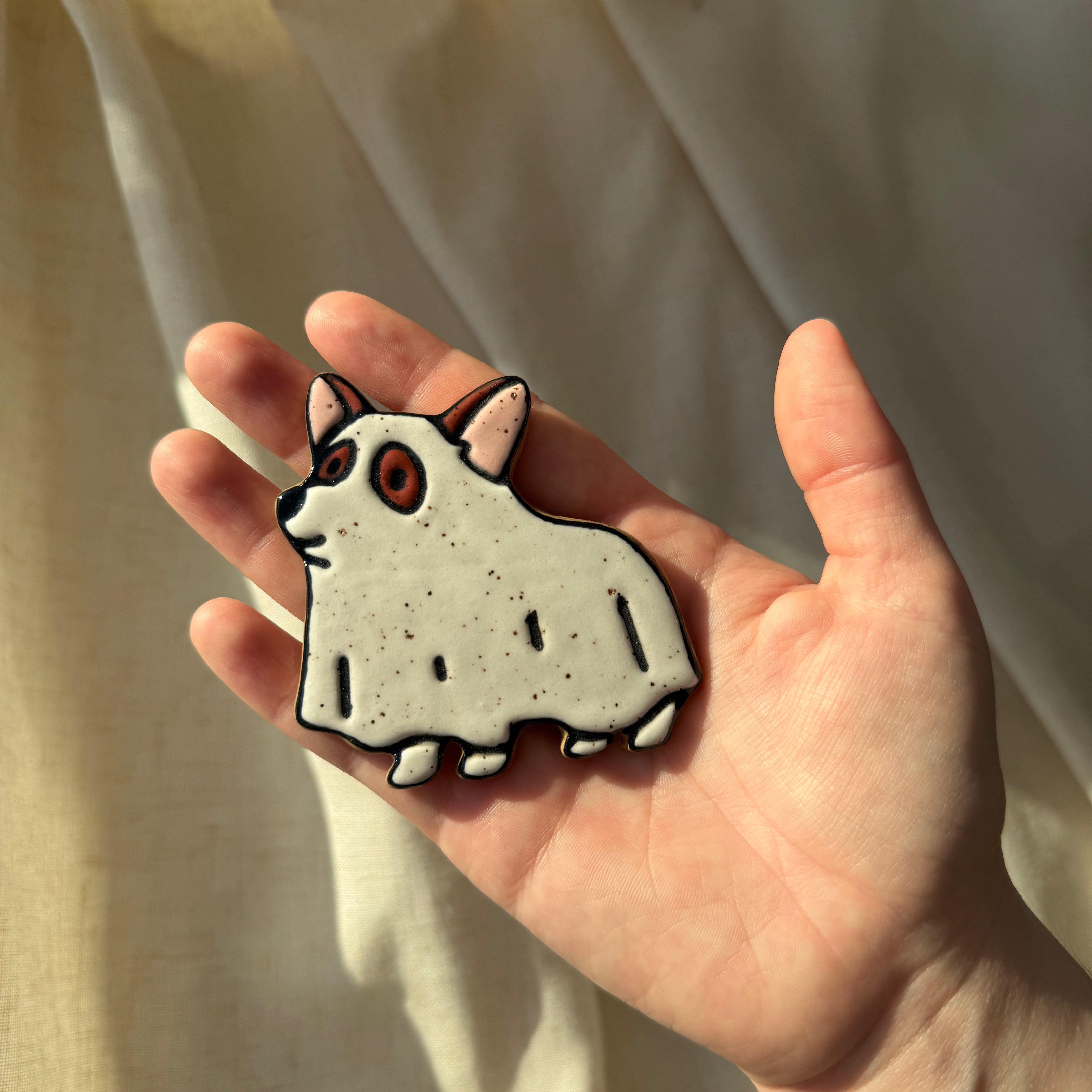 A hand holding a small ceramic magnet shaped and handpainted as a corgi with a ghost sheet draped over the dog, set against draped cream fabric.