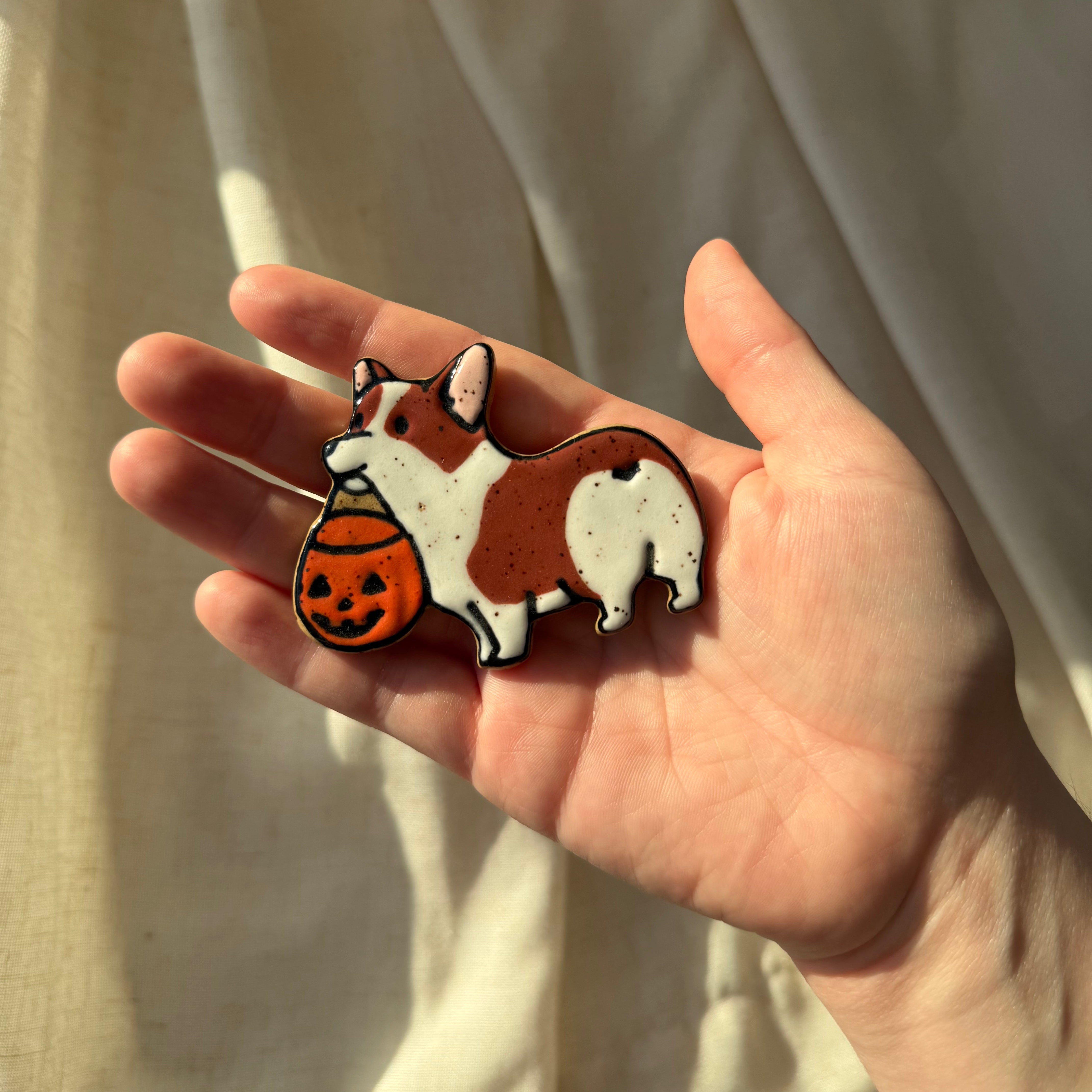A hand holding a small ceramic magnet shaped and painted like a brown and white corgi holding an orange pumpkin bucket, set against a background of draped cream fabric.